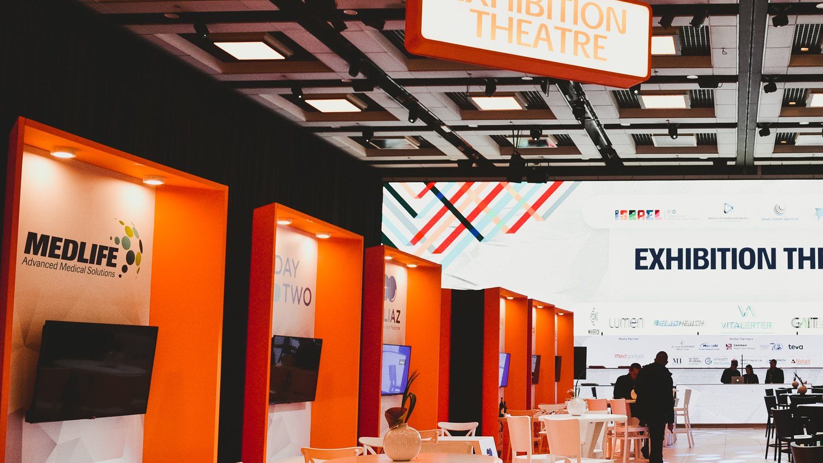 A receding row of orange exhibition bays with wall-mounted screens, an Exhibition Theatre sign hanging above.