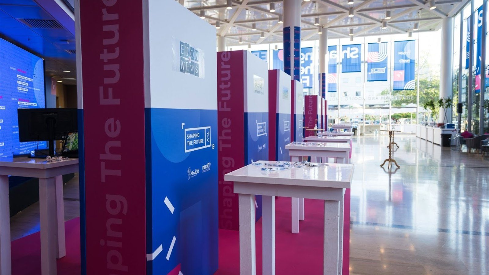 A row of blue and magenta totems receding across a magenta floor in a glazed atrium.