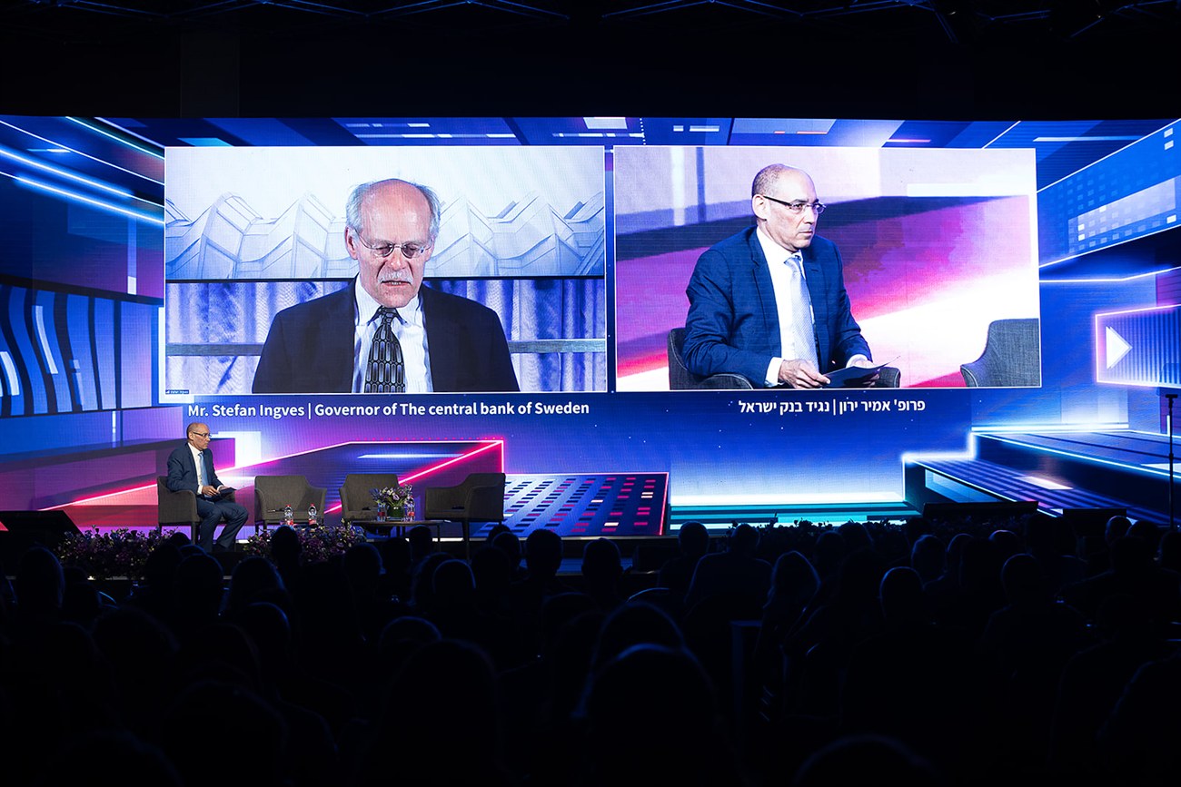 Two speakers shown side by side on the full-width stage wall, captioned as the Governor of the central bank of Sweden and the Governor of the Bank of Israel, the audience in silhouette below.