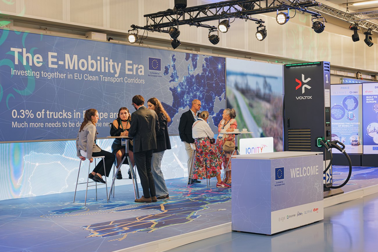 The E-Mobility Era exhibition in use: a lounge in front of a wall-wide map of European transport corridors, a VOLTIX charger and a welcome desk on a lit map floor.