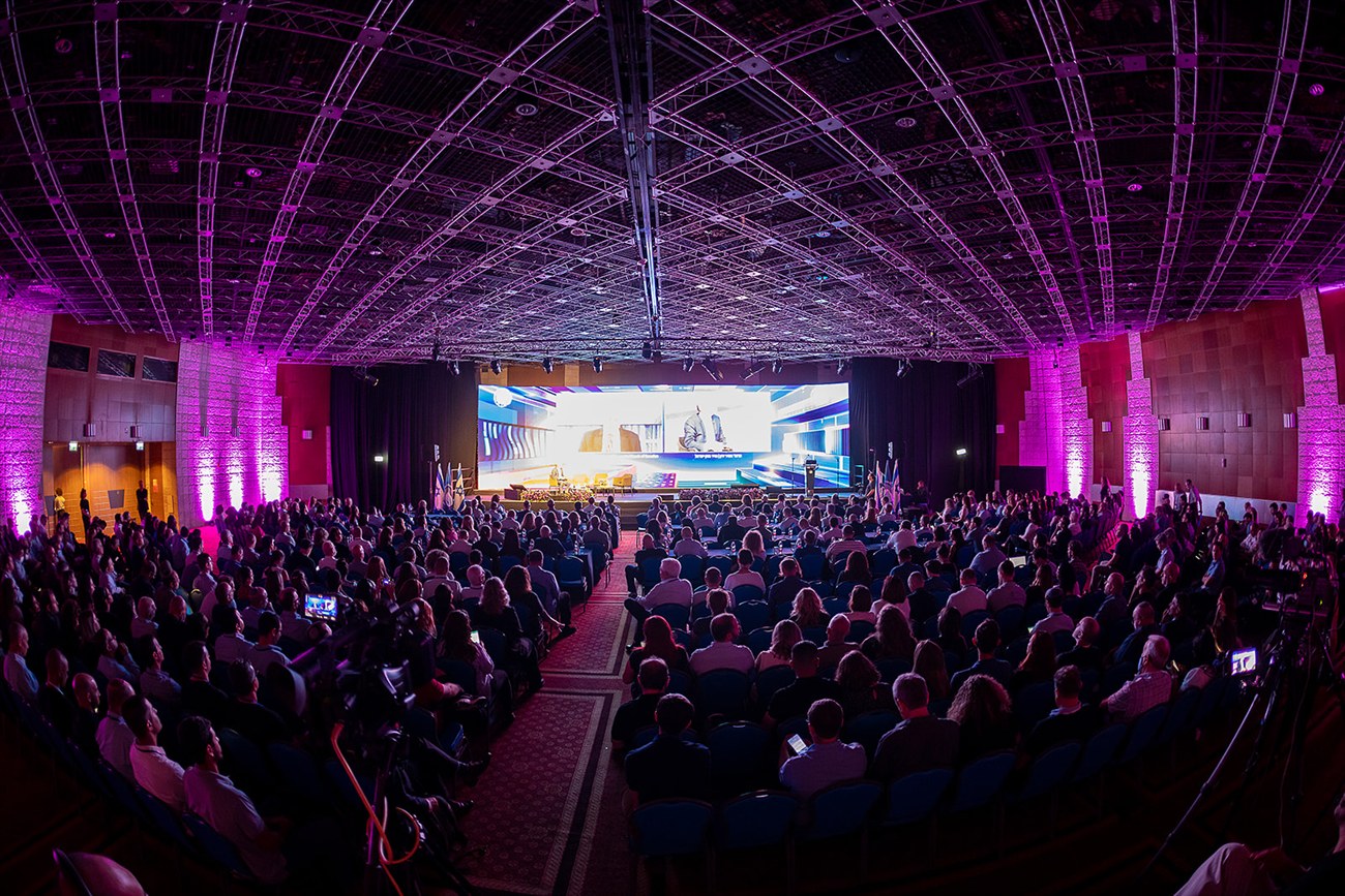 A full conference hall seen from the back, magenta uplighting along the walls and a curved lighting truss overhead, the stage screen small in the distance.