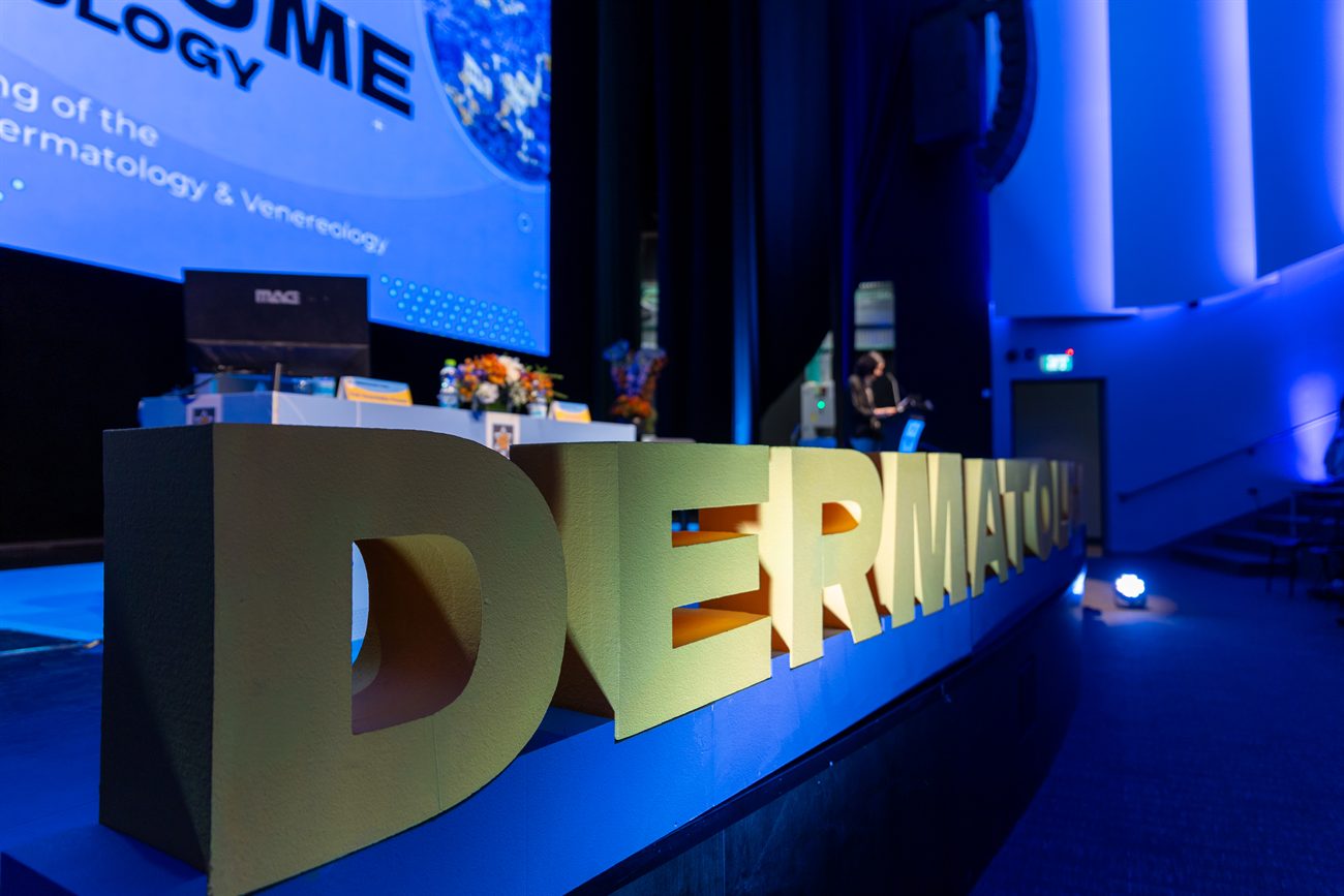 Freestanding gold letters spelling the conference name across the front of a stage lit blue, a panel table and a wide screen behind them.