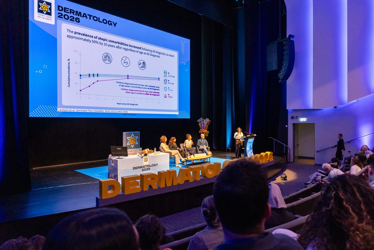 The stage seen from within the audience, the screen and the panel table beyond rows of seated delegates.