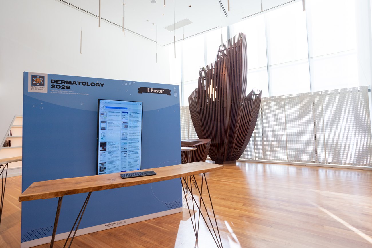 An e-poster station in a hall lit by daylight: a printed panel carrying the conference name, a screen and a long wooden table.