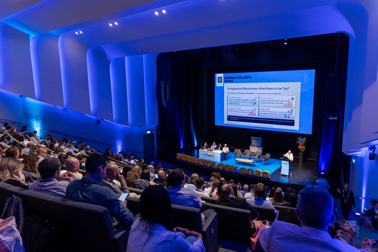 The main hall from the back rows in session, the full stage set and the screen at the far end of a curved, blue-lit auditorium.