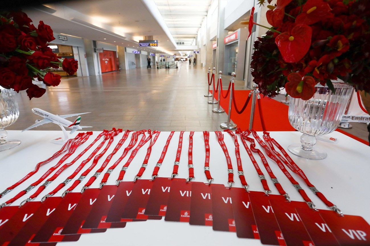 VIP lanyards and cards laid out in a row on a white table, red roses beside them, a red carpet running away into the terminal.