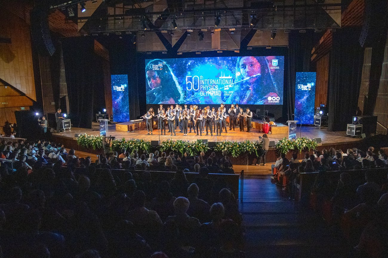 The closing ceremony seen from the back of a full hall, a wind orchestra on stage beneath a wide screen.