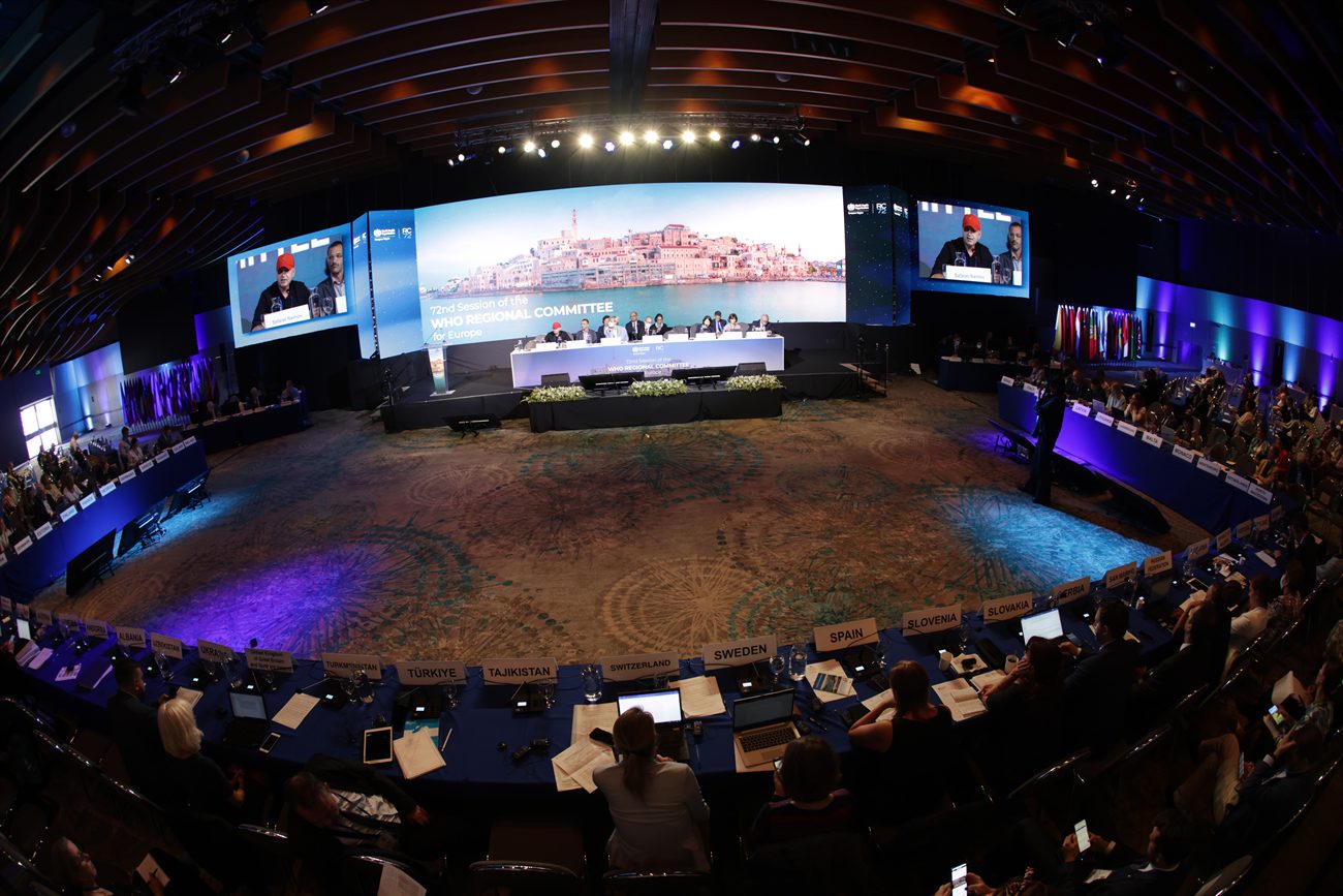 The assembly in session seen from above, delegate desks curving in a full horseshoe around an open floor, a panoramic screen wall behind the platform and country nameplates along the near row.
