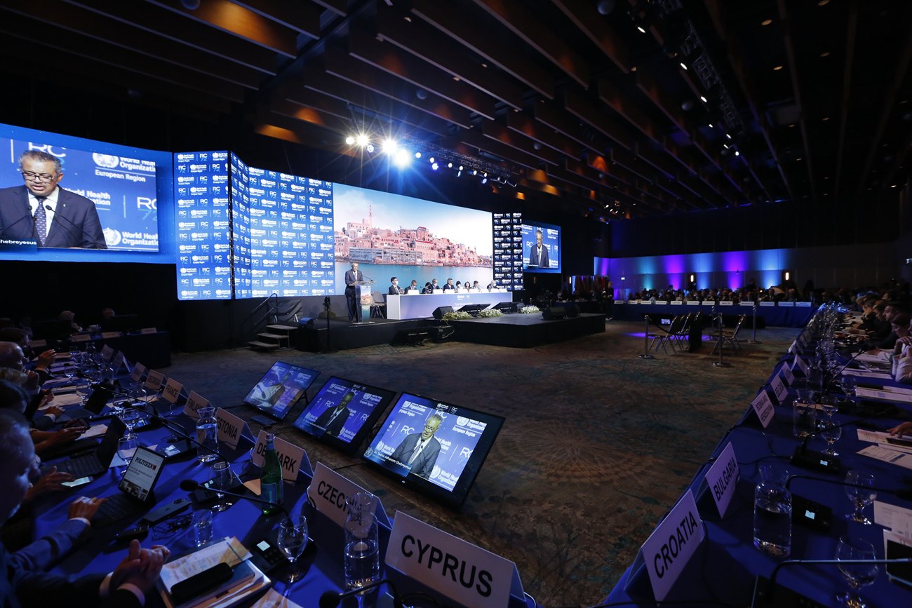 The assembly in session from the delegate floor: the stage and a wall of screens beyond, desk monitors and country nameplates in the foreground.