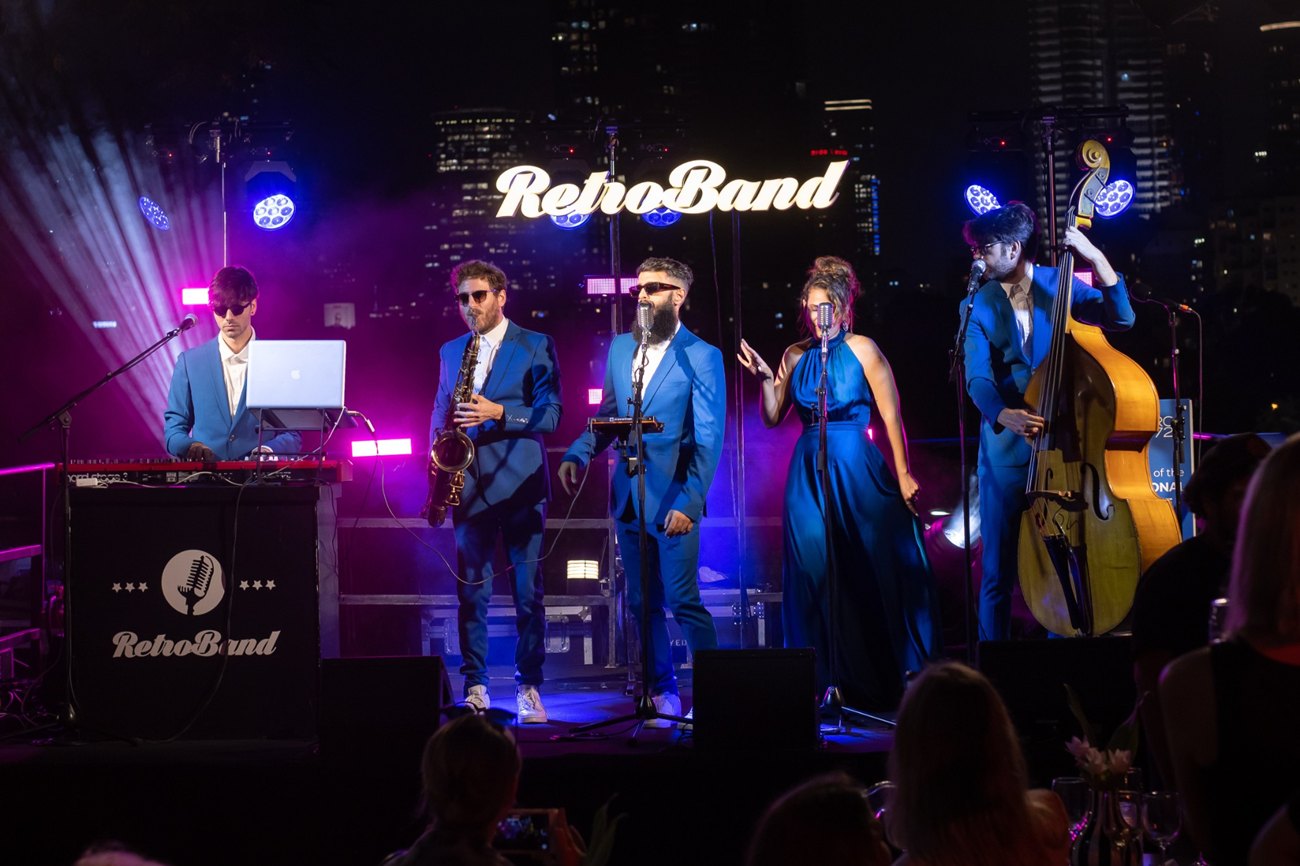 A five-piece band in blue suits playing on an outdoor stage at night, the city's towers lit behind them.